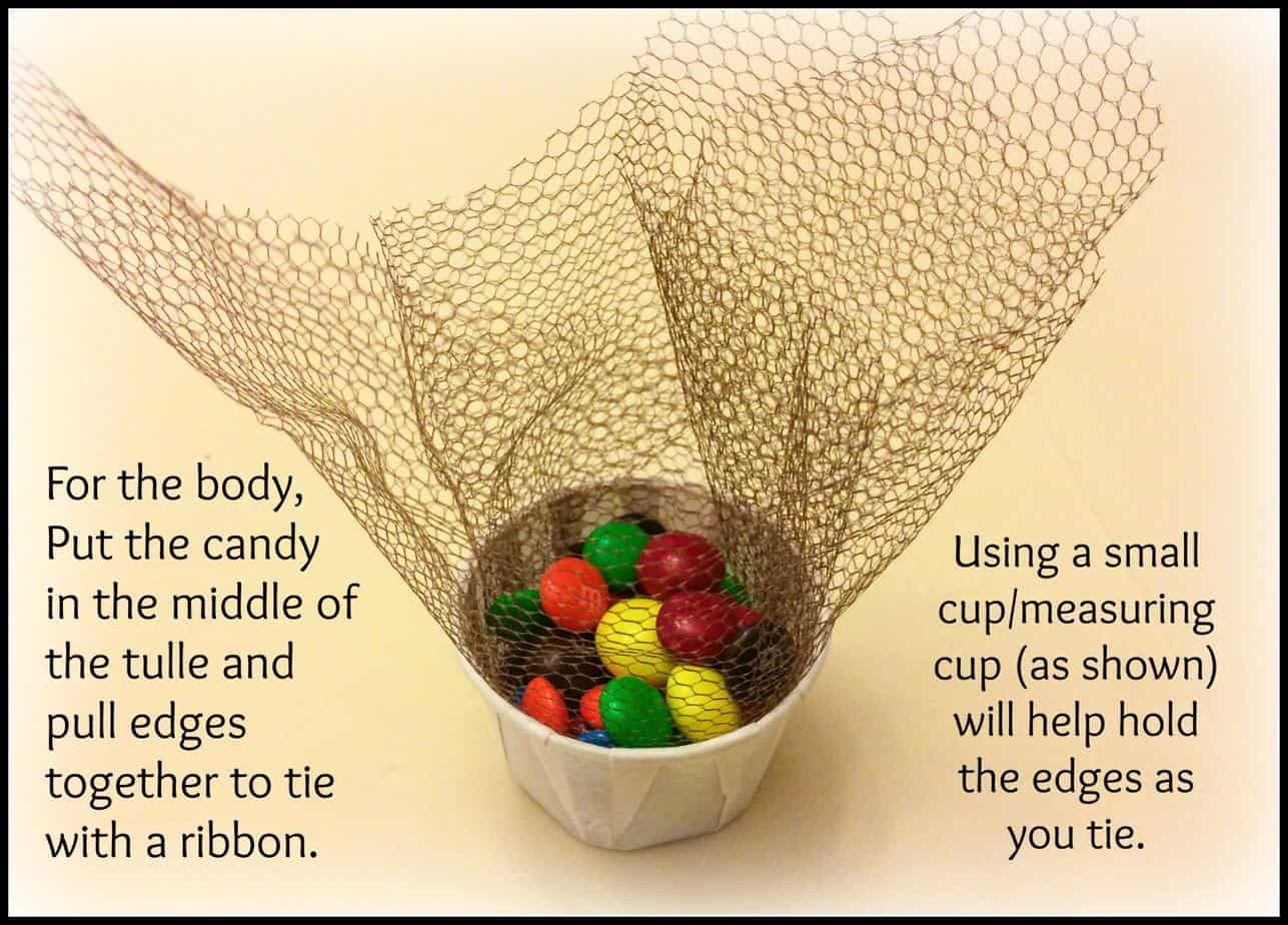 M&M's in tulle. Says "For the body, put the candy in the middle of the tulle and pull edges together to tie with a ribbon. Using a small cup/measuring cup (as shown) will help hold the edges as you tie.